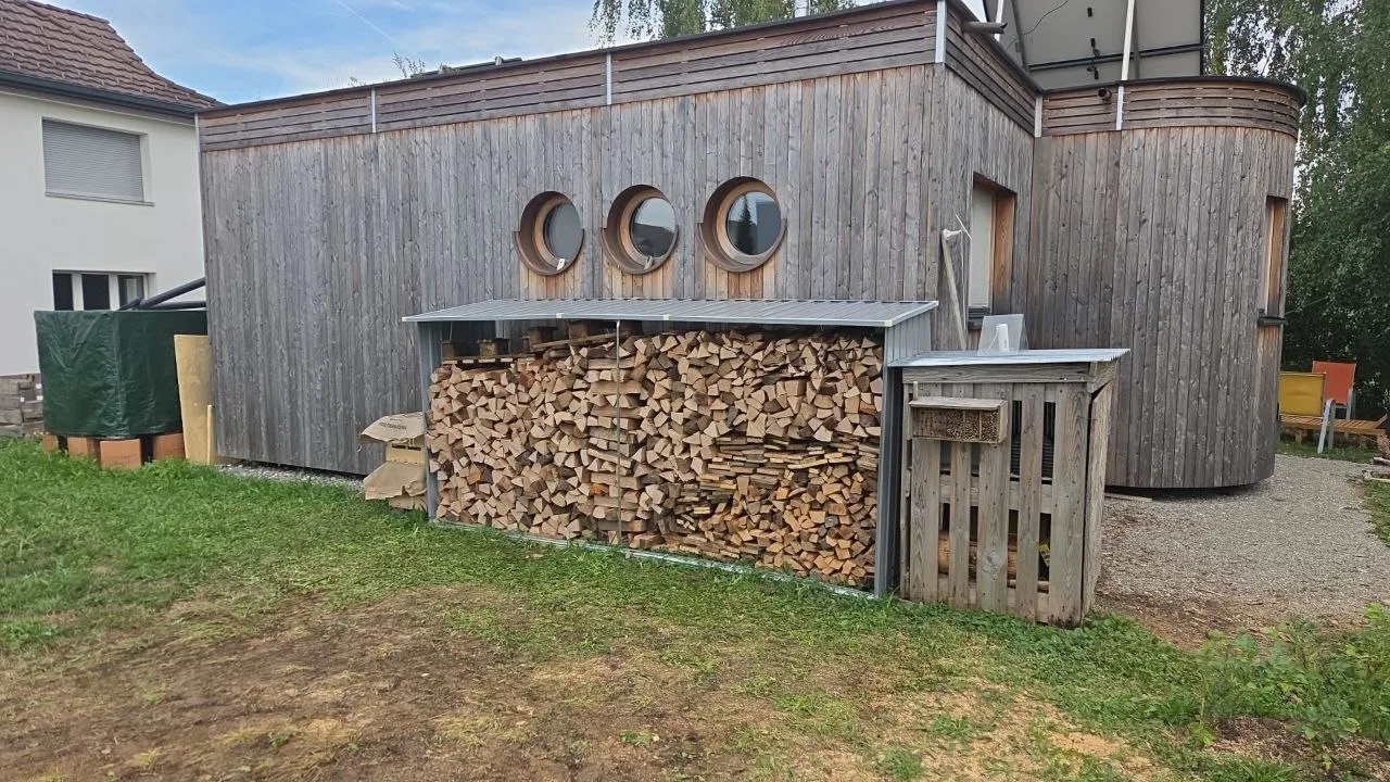 Holzverkleidetes Gebäude mit drei runden Fenstern und einem Unterstand voller gestapeltem Brennholz.