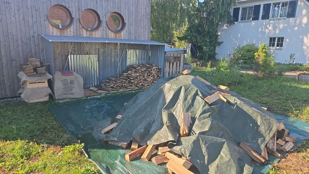 Holzstapel und Holzhaufen unter Planen liegen neben einem Holzschuppen mit runden Fenstern im Garten.