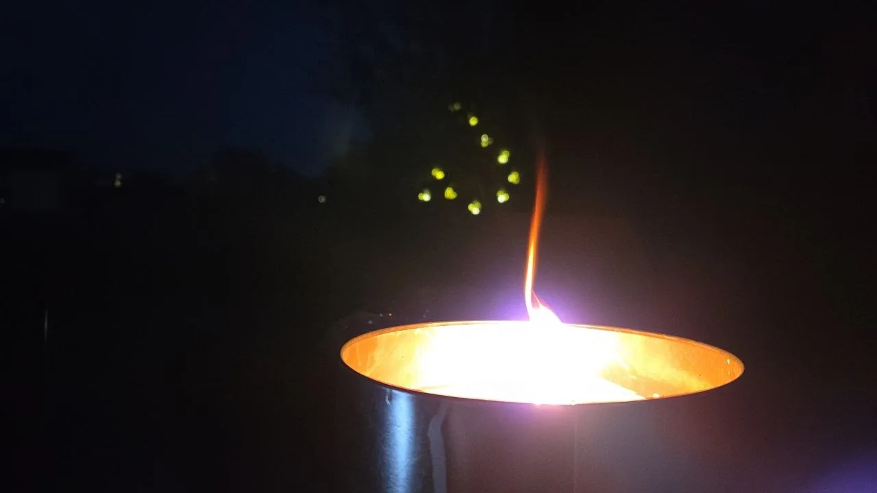 Flamme steigt aus einem Metallbecher in dunkler Umgebung auf, im Hintergrund sind gelbe Lichtpunkte zu sehen.