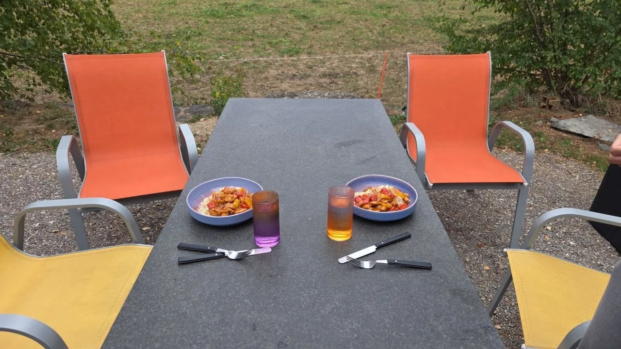 Auf einem Gartentisch stehen zwei Teller mit Essen, zwei Gläser und Besteck, umgeben von vier Stühlen.