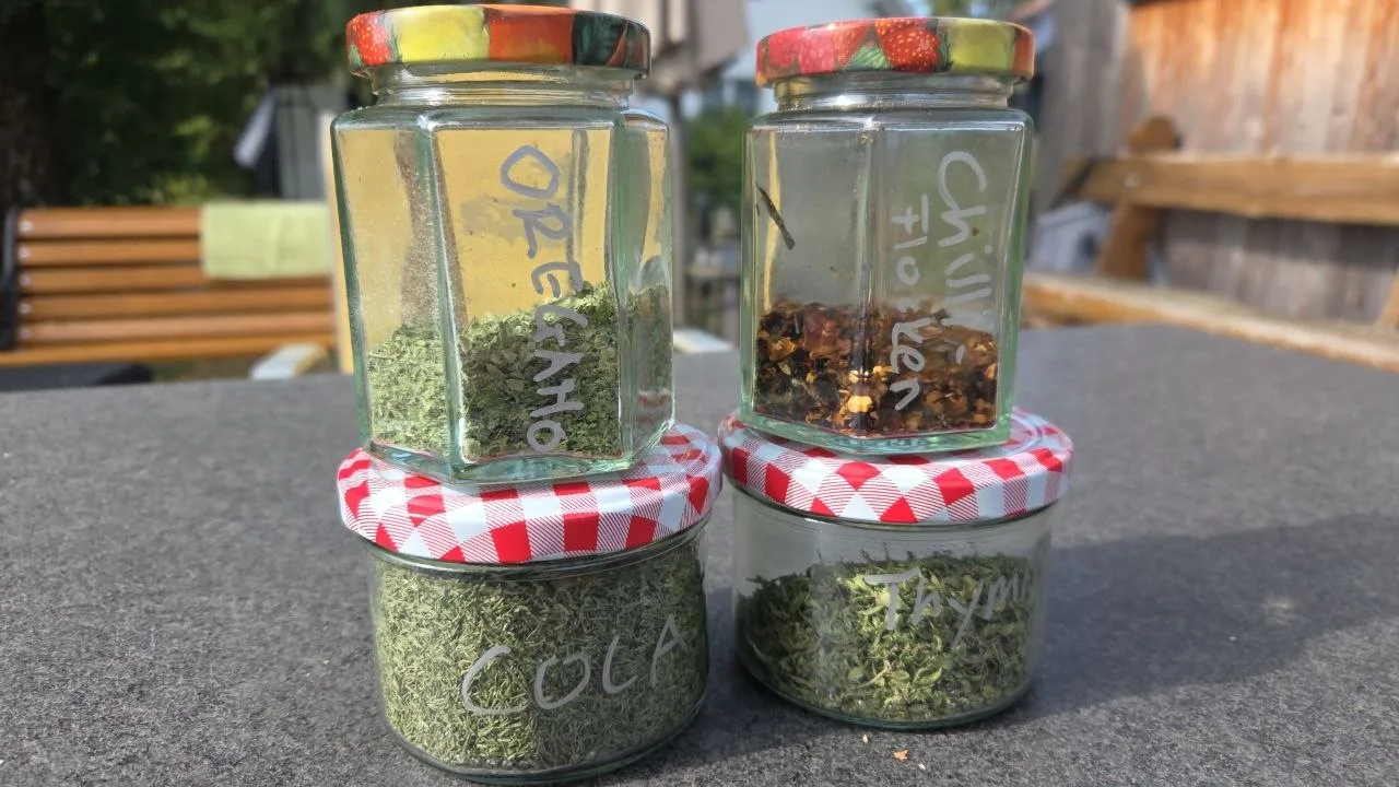 Vier beschriftete Gläser mit getrockneten Kräutern und Chiliflocken stehen auf einem Tisch im Freien.